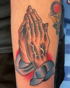 Traditional praying hands tattoo with red background on forearm by San Diego tattoo shop